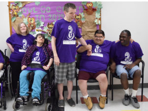 A group of people with varying abilities is seated in a classroom. The individual in the center is standing. They are wearing matching purple t-shirts.
